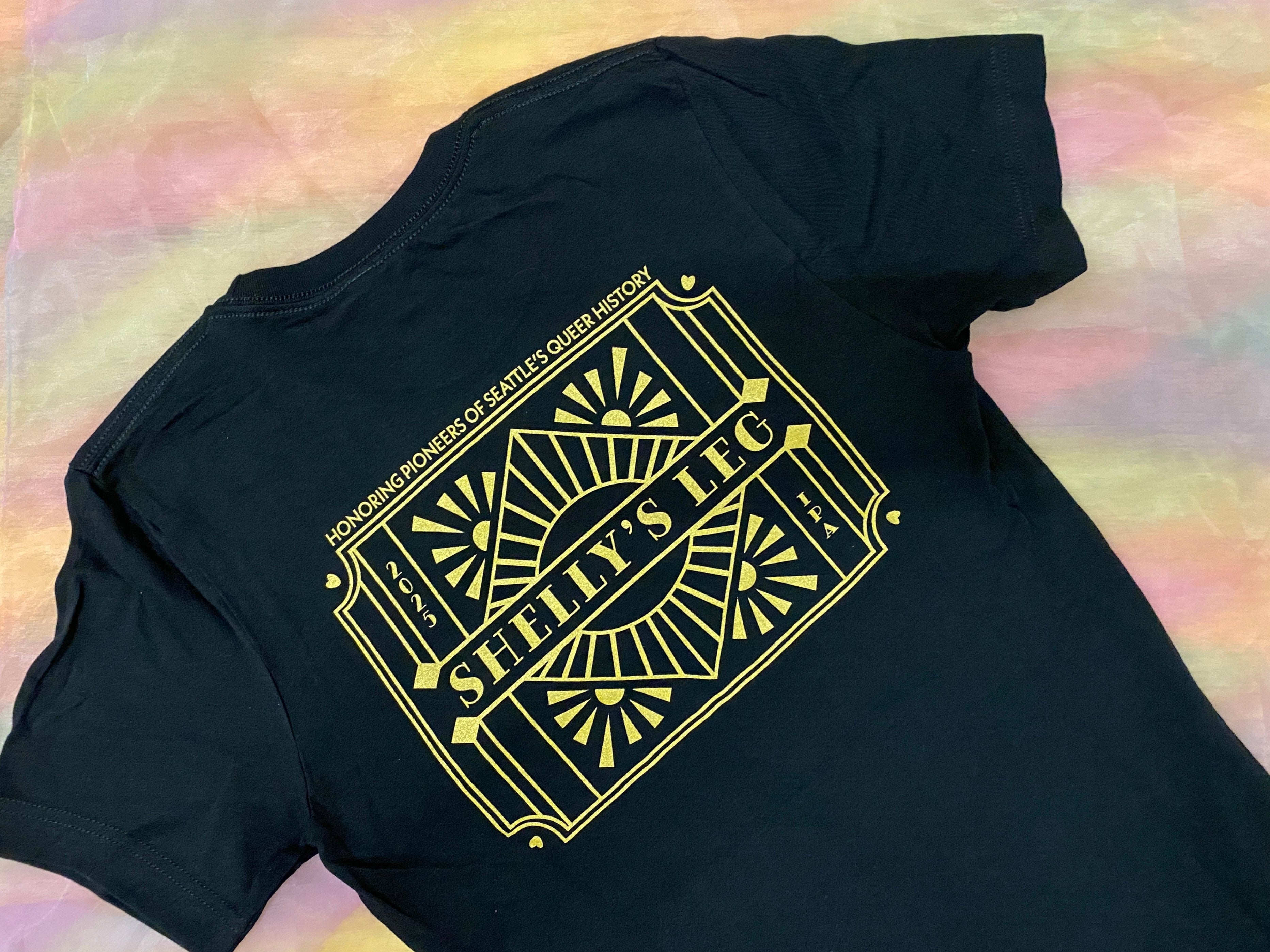 black shirt with an art deco style design that reads: honoring pioneers of seattle's queer history, shelly's leg IPA, 2025
