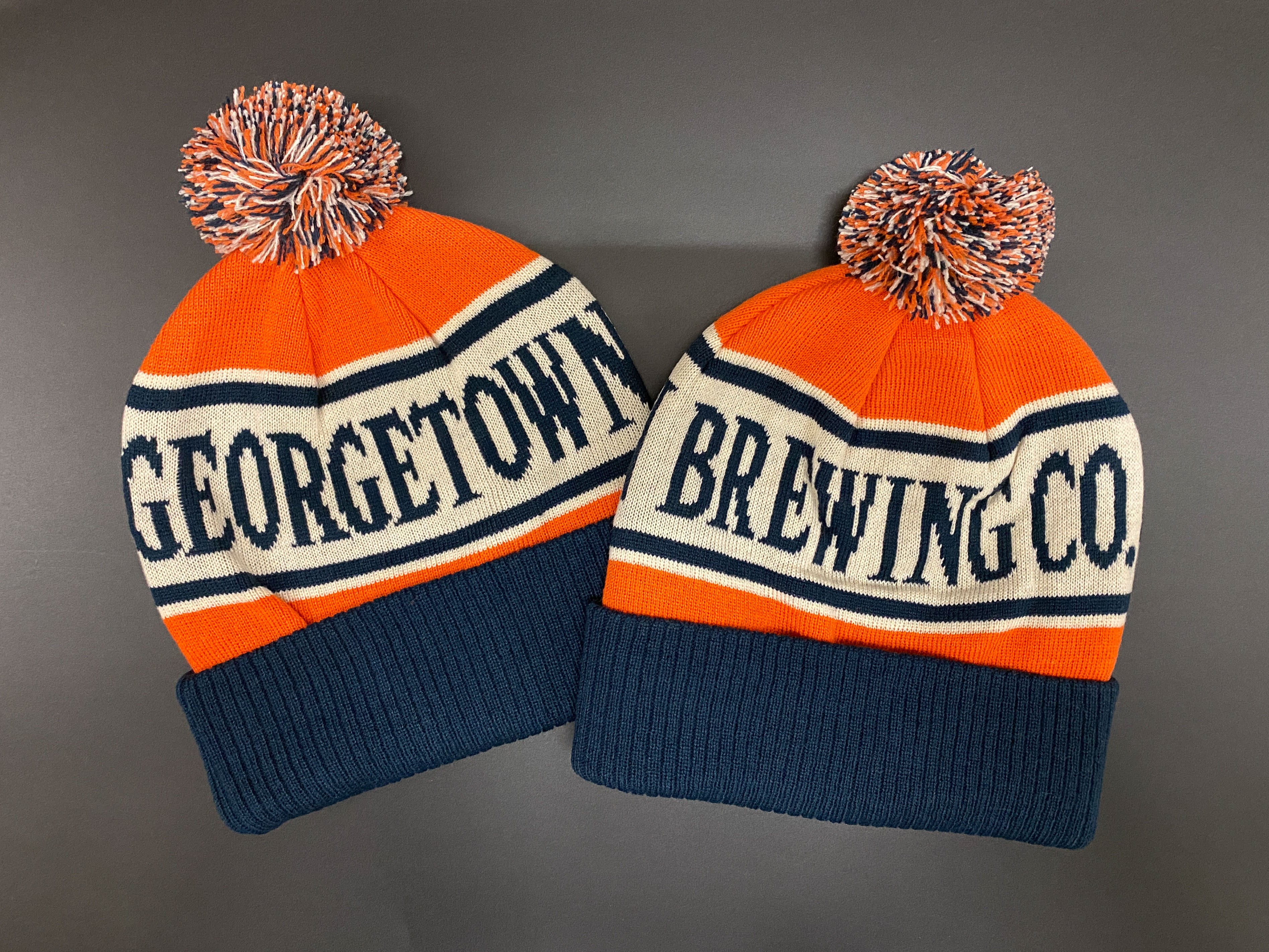Two Georgetown Brewing Co. beanies with pom-poms on a gray background. The colors are orange, blue and cream that are supposed to be Bodhizafa colors but will undoubtedly be mistaken for Bears or Broncos colors. 