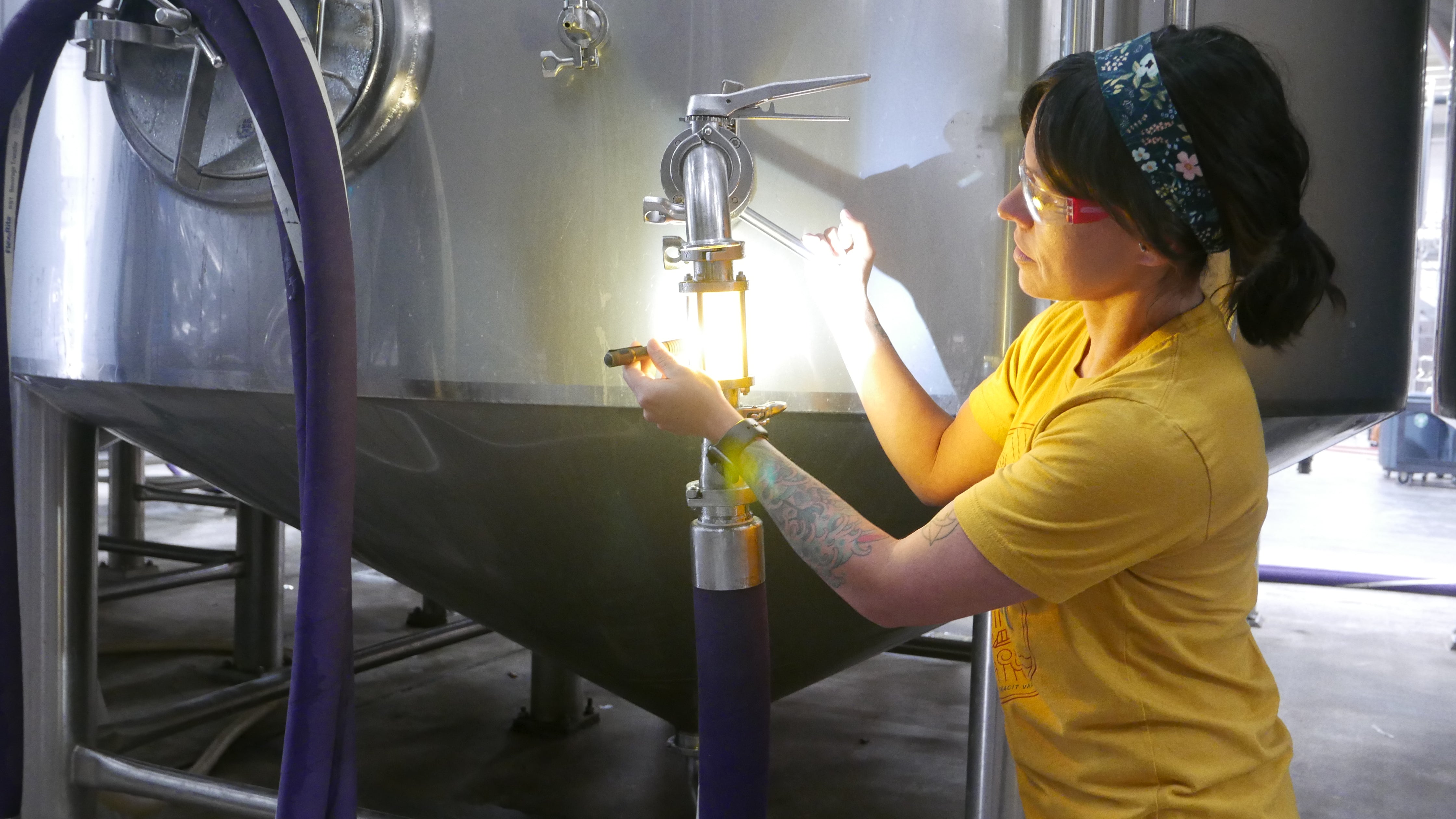 A woman with dark hair uses a flashlight to look at a sightglass connected to a beer tank