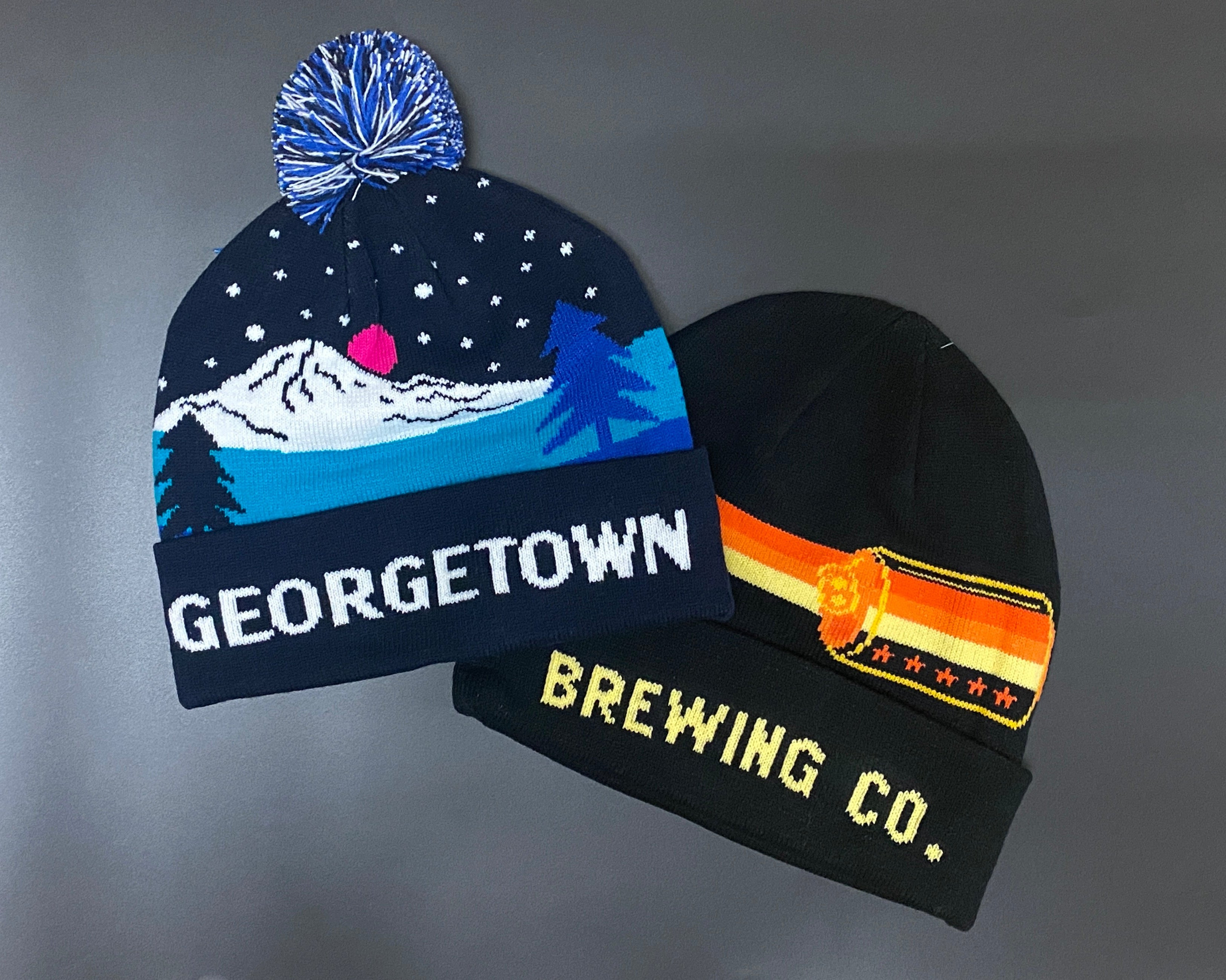 Two knit  beanie hats laid flat on a gray background. The left hat is navy with a mountain-and-sky graphic and the word GEORGETOWN in white. The right is a yellow and orange can design that says Brewing Co. 