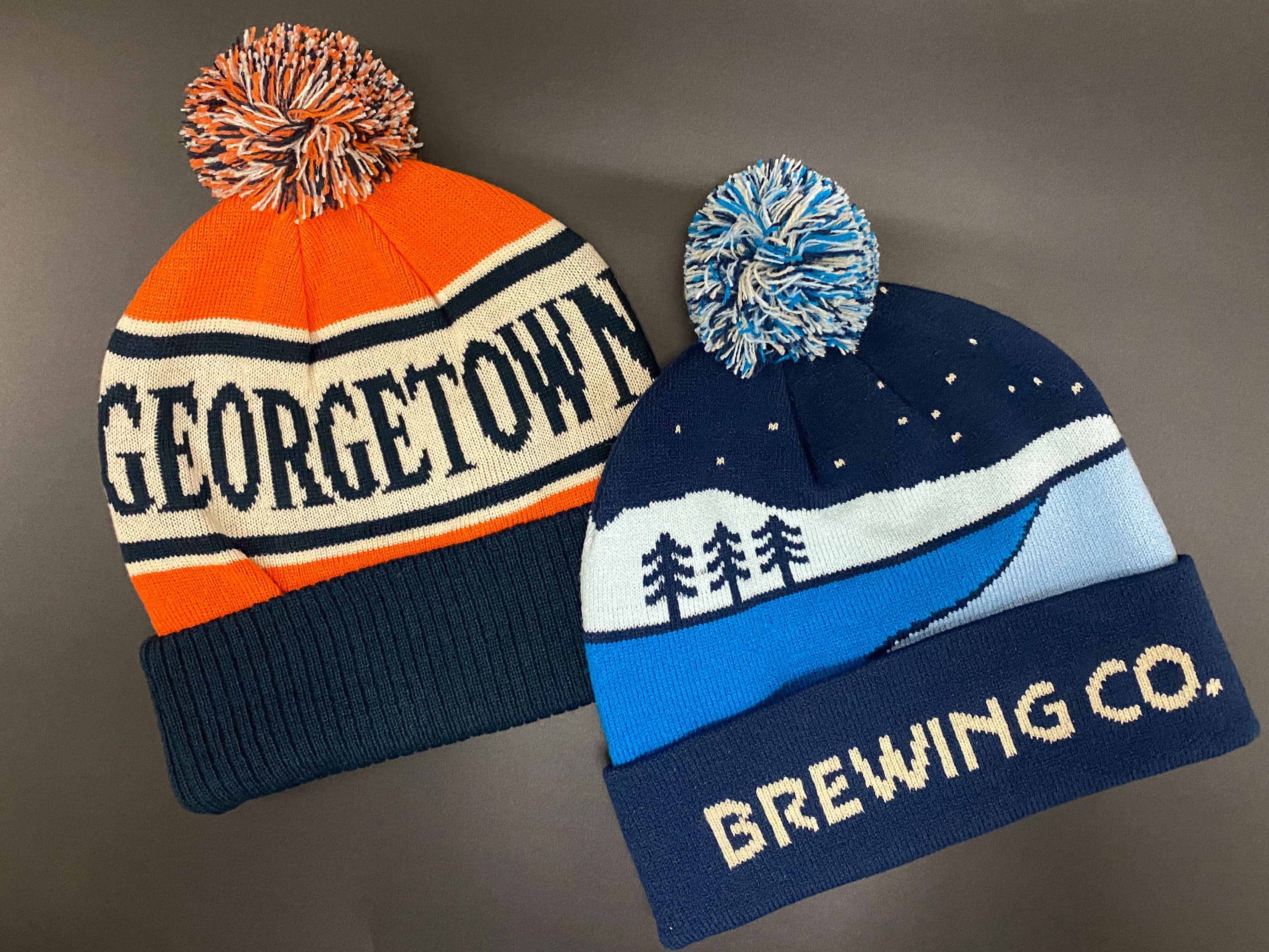 Two knit hats with pom-poms, one labeled 'Georgetown' and the other 'Brewing Co.', on a gray background.