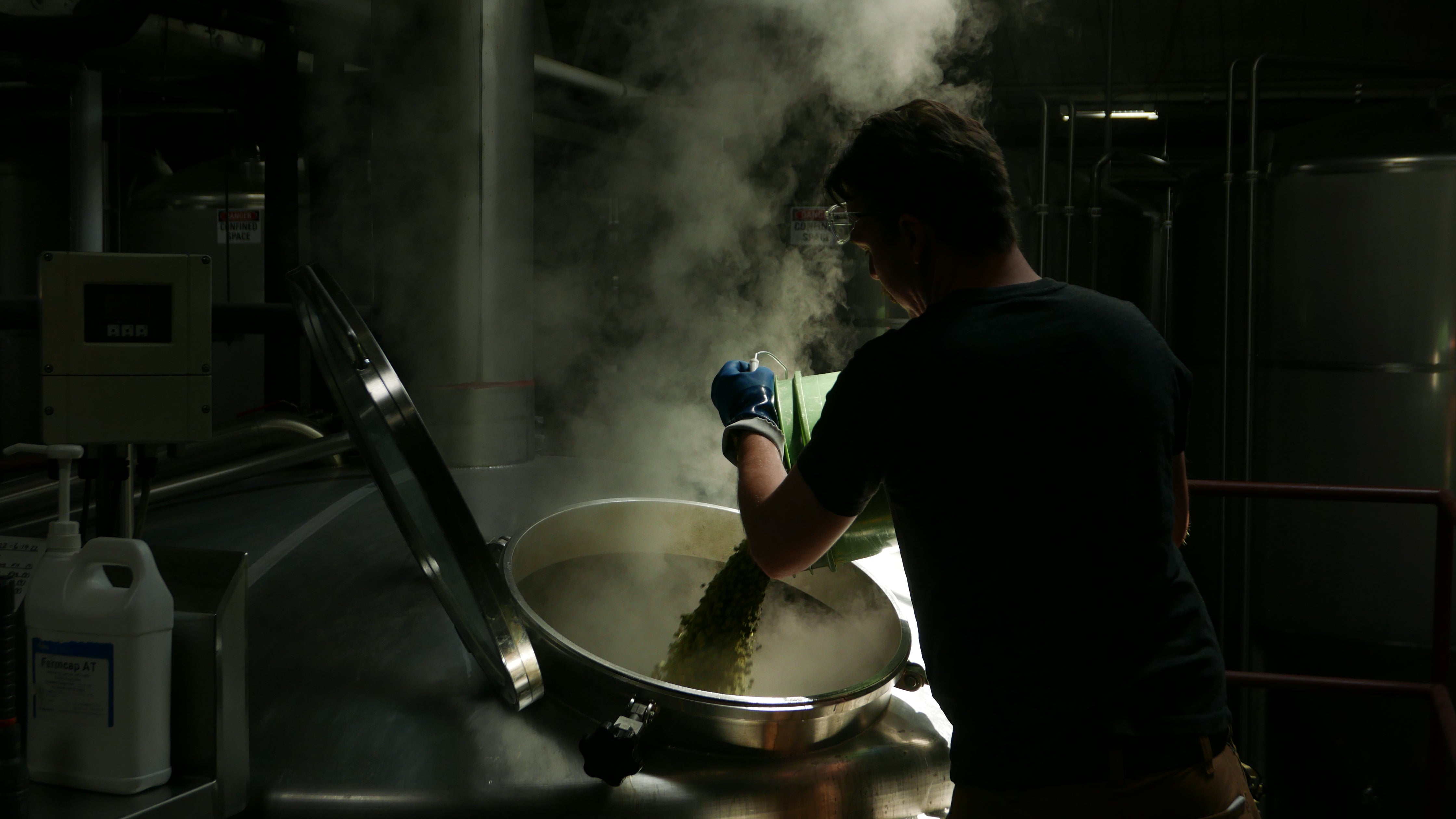 the sun shines own on to a brew tank while a brewer adds hops to the tank. The steam is illuminated and it looks very moody. 
