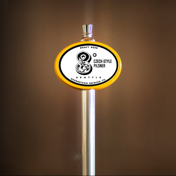 A close-up of a beer tap handle with an oval white, black, and yellow logo that reads '8 degrees Czech-Style Pilsner set against a dark brown blurred background.