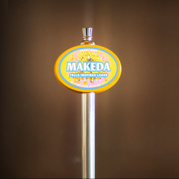 A close-up of a Makeda beer tap handle with a round yellow, blue, and white logo on a silver metal post against a dark brown blurred background.