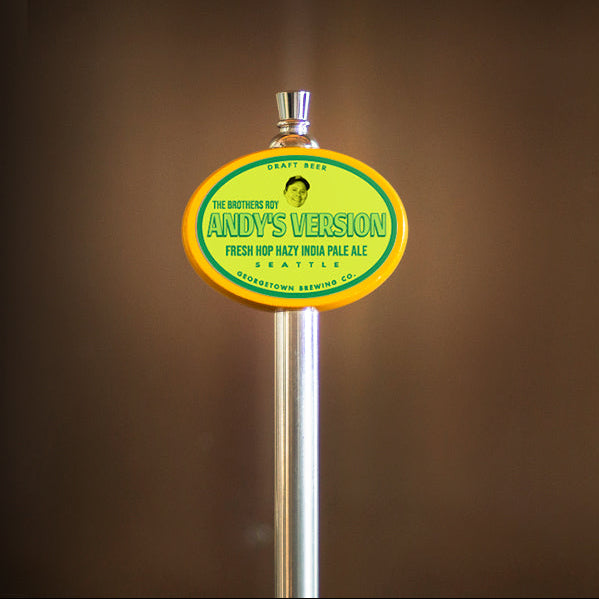 A close-up of a beer tap handle with a round yellow and green label reading 'Andy's Version Fresh Hop Hazy IPA Pale Ale' against a dark brown blurred background.