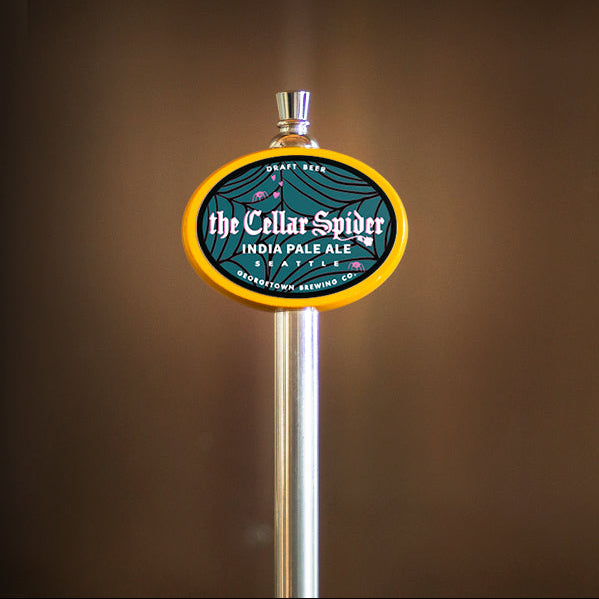 A close-up of a beer tap handle with an oval label reading 'The Cellar Spider India Pale Ale' on a blurred brown background.
