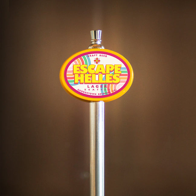 Colorful 'Escape Helles' beer tap handle against a brown background