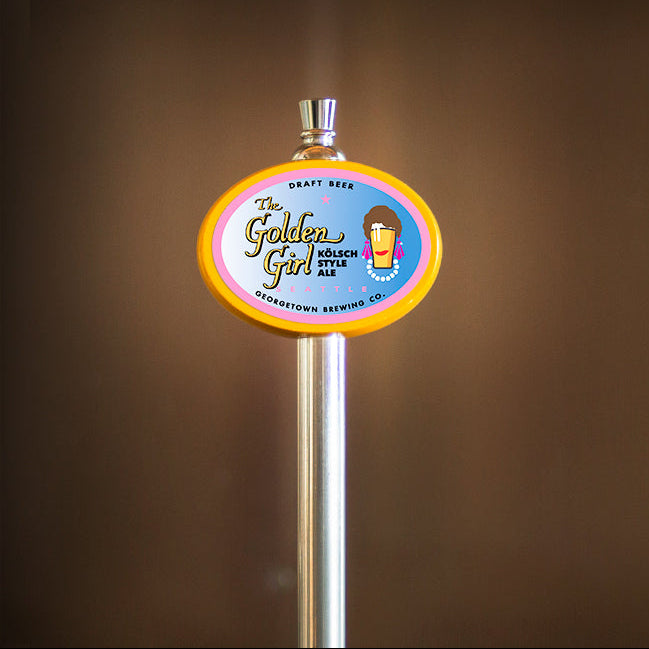 Beer tap handle with text reading The Golden Girl and a pint of beer wearing earrings, pearl necklace, has a brown buffount hairdo and lipstick to mimmick a golden girl from the tv series. 
