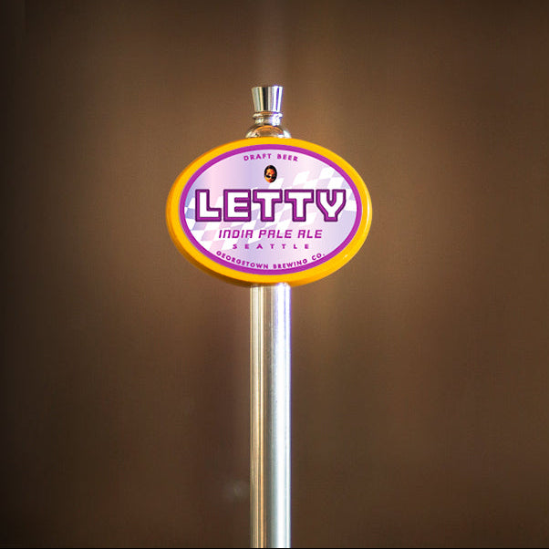 A close-up of a beer tap handle with a round Letty India Pale Ale logo in purple, pink, and yellow, mounted on a silver metal post against a dark brown blurred background.