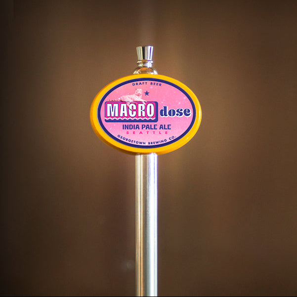 A close-up of a beer tap handle with a circular logo reading 'MACRO dose' and 'INDIA PALE ALE' on a metallic pole against a blurred brown background.