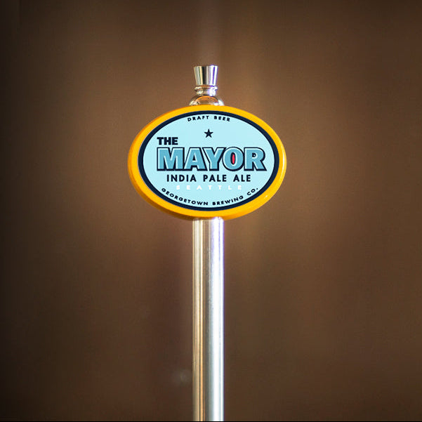 A close-up of a beer tap handle with an oval sign reading 'THE MAYOR INDIA PALE ALE' in blue and white with a yellow border, set against a dark brown blurred background.