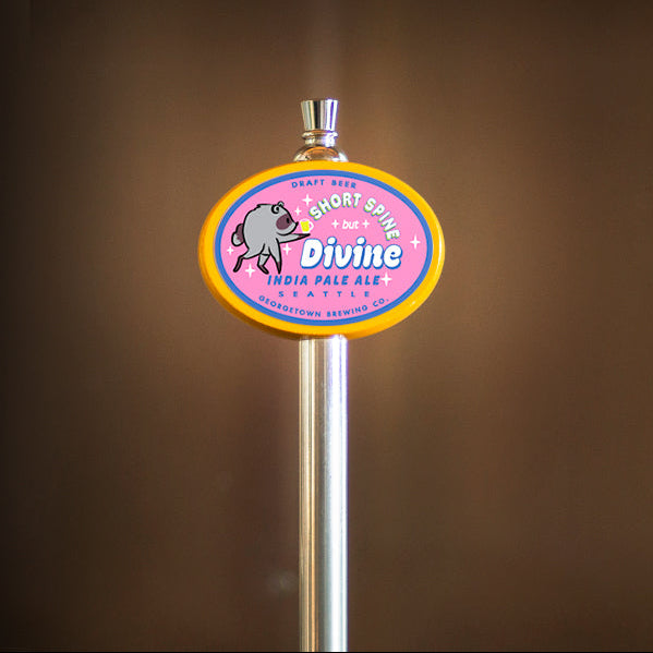 A close-up of a beer tap handle for Mount Sweet Divine India Pale Ale, featuring a purple and yellow oval label with a Jimothy graphic, set against a dark brown blurred background.