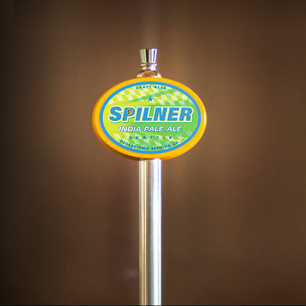 A close-up of a beer tap handle featuring a circular Spilner India Pale Ale logo with a yellow border, green and blue label, and silver metal stem against a dark brown blurred background.