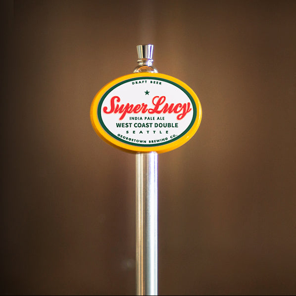 A close-up of a beer tap handle with a round 'Super Lucy' logo sign, set against a dark brown blurred background.