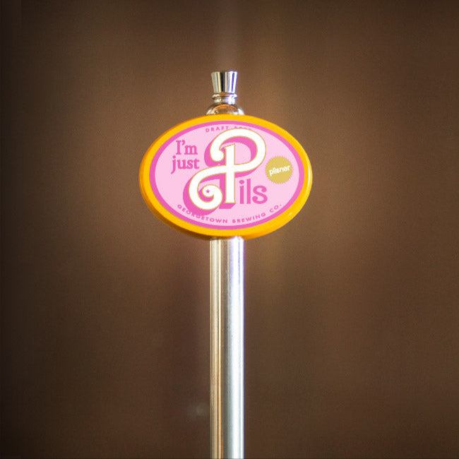 Beer tap handle with a pink and yellow I'm Just Pils label on a brown background